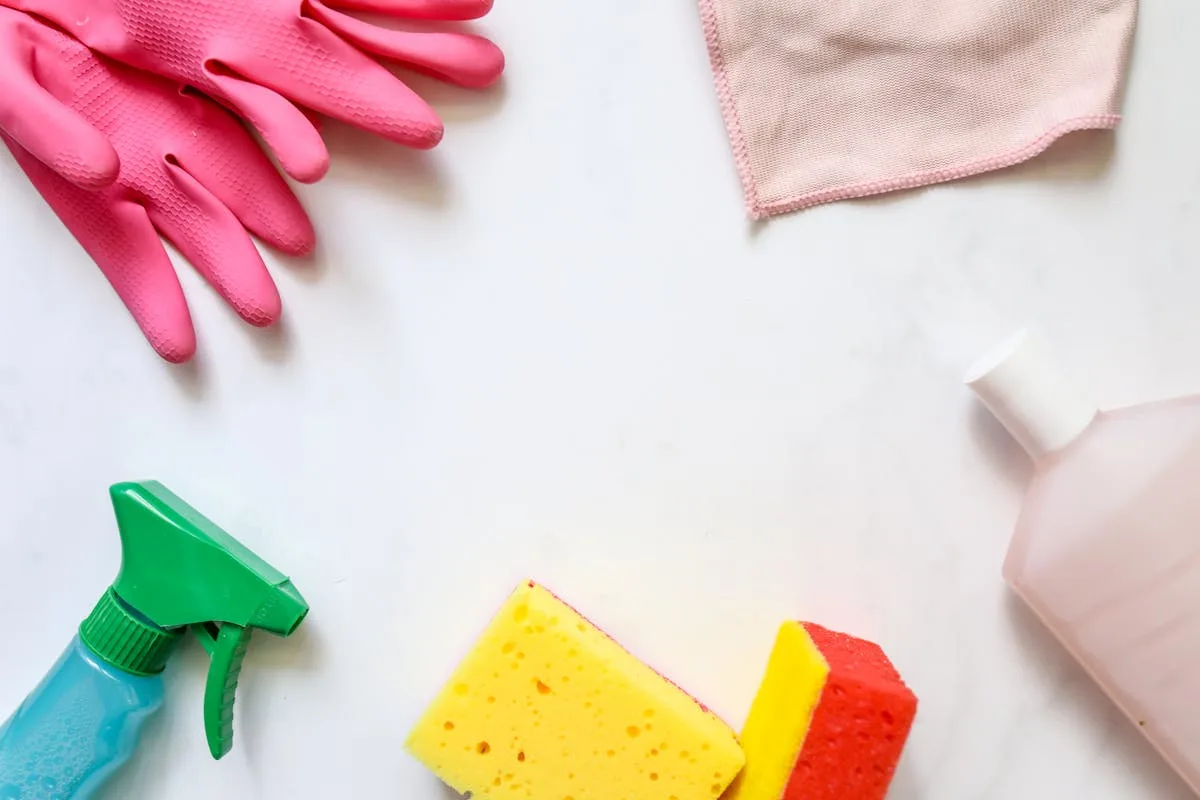 Flat lay of cleaning supplies with pink gloves, sponges, a cloth, and a spray bottle on white marble