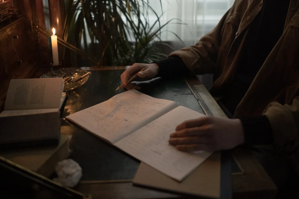 Person in earth-toned jacket writing in a journal at a wooden desk lit by candlelight