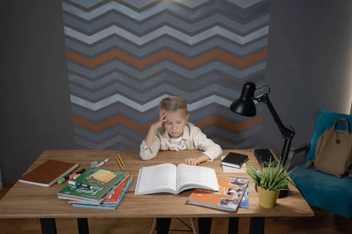 Child working through schoolbooks at a wooden desk with a lamp and stacked workbooks