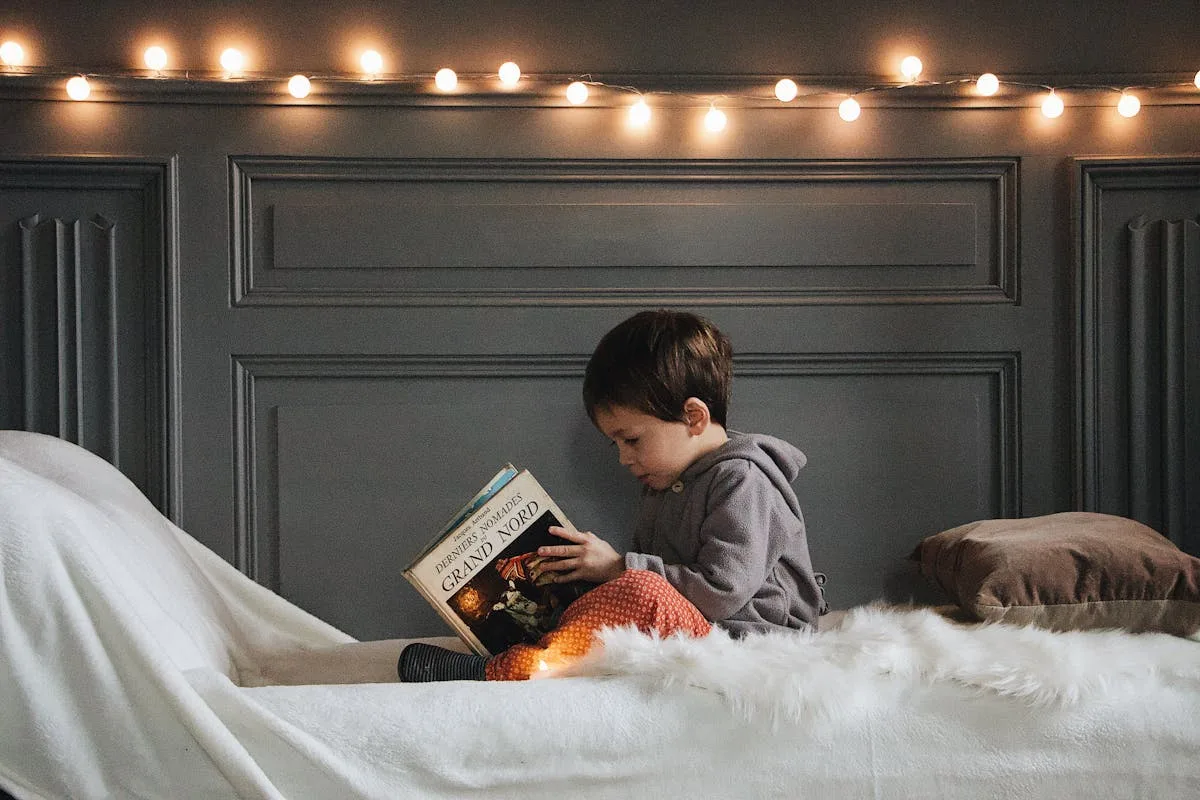 Young child reading a book on a bed under warm string lights