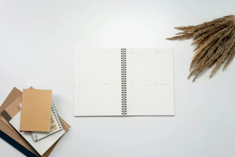Open weekly planner notebook on a white desk beside a stack of kraft notebooks