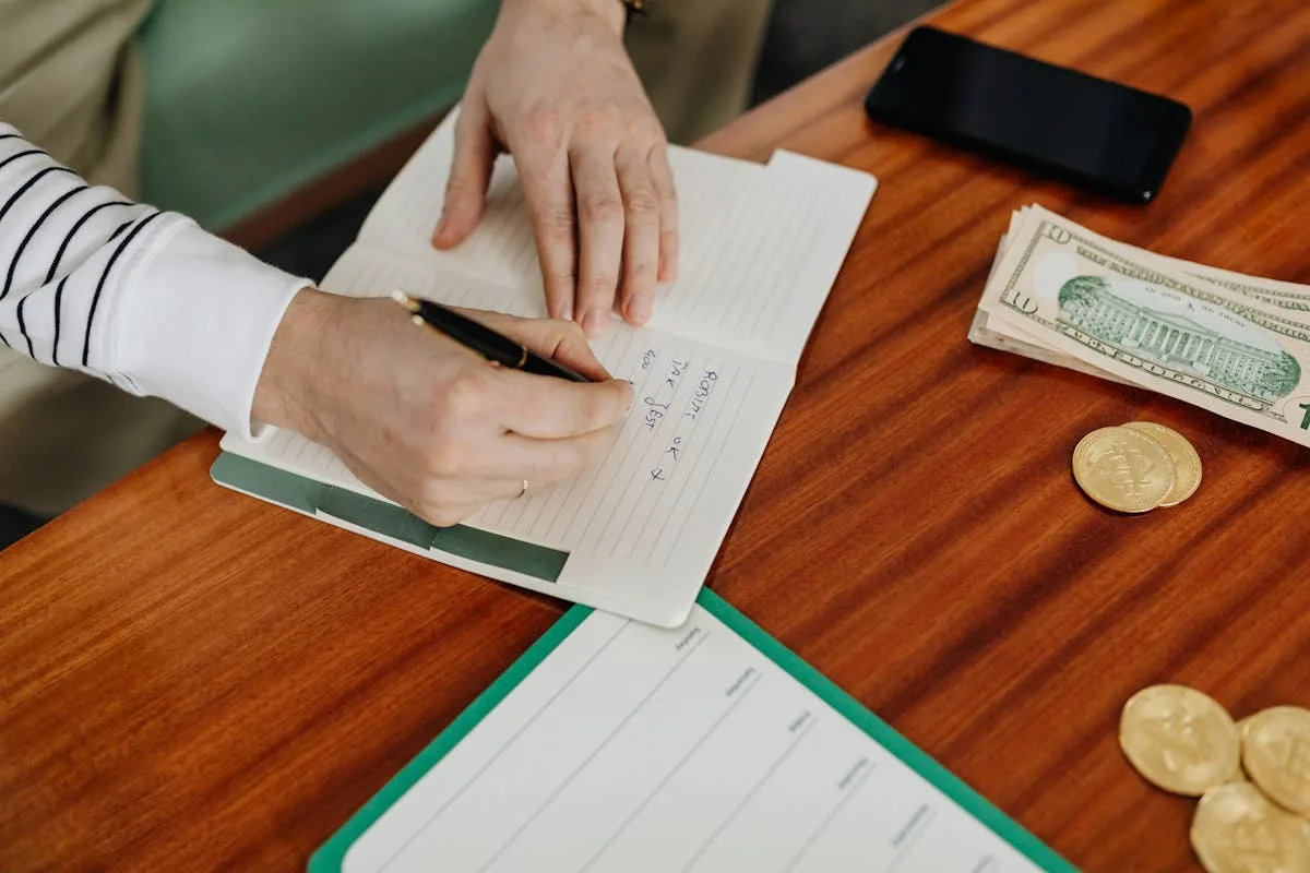 Hands writing a wedding budget in a notebook with cash and coins on a wood table