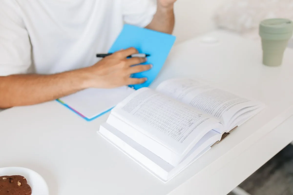 Student at a white desk writing notes beside an open textbook and a coffee cup