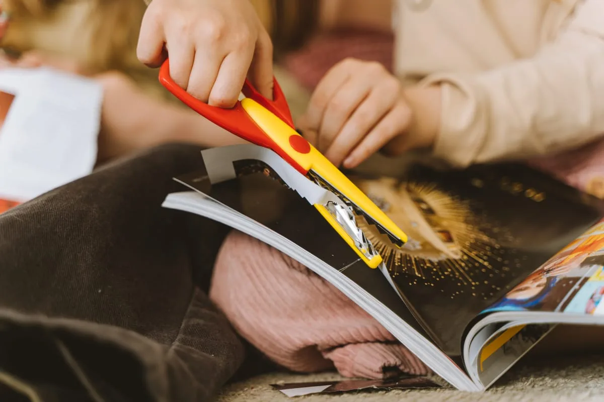 Hands cutting an image out of an open magazine with red and yellow scissors