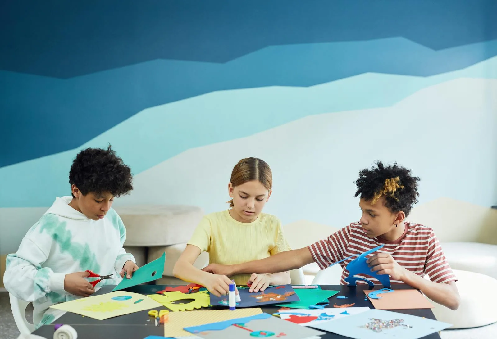 Three friends sitting at a table together cutting and gluing magazine pieces in a softly lit room