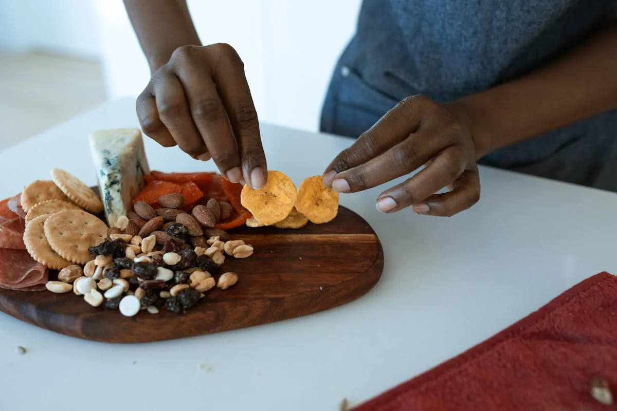 Hands assembling a small snack board with crackers, nuts, dried fruit and cheese on a wooden serving board