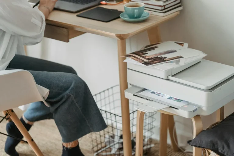 A woman at a small wooden desk with a home printer feeding printed pages from below