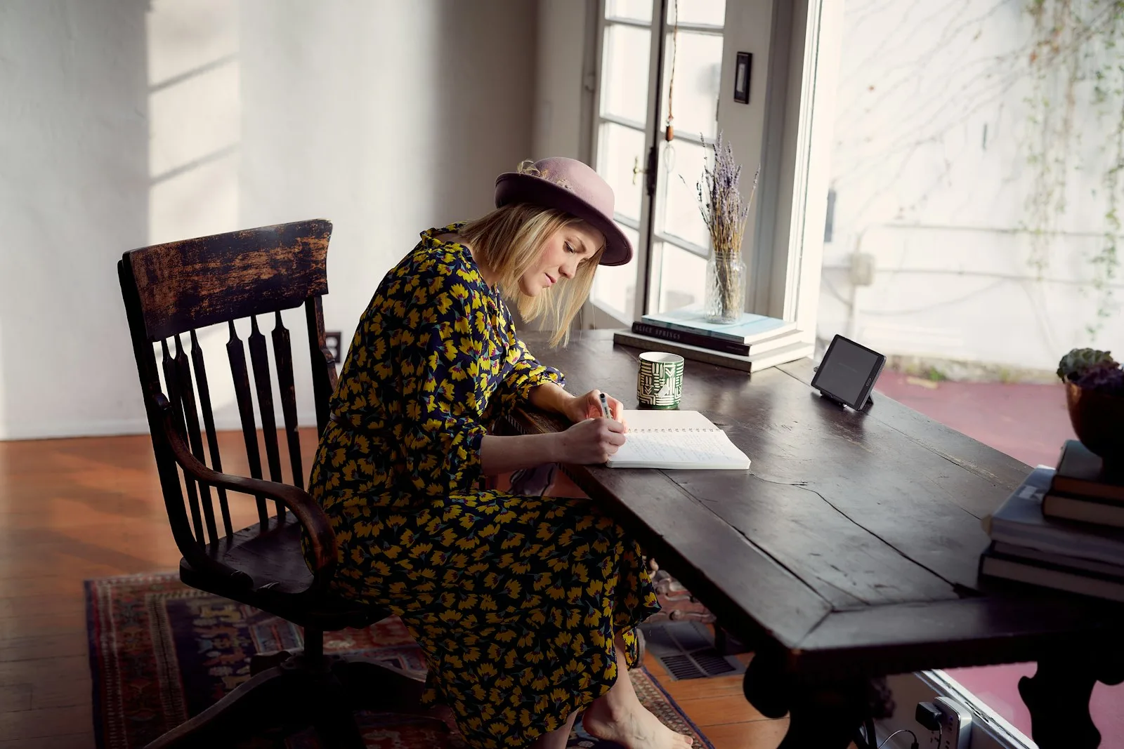 Woman writing in a journal at a wooden table beside a window in warm afternoon light