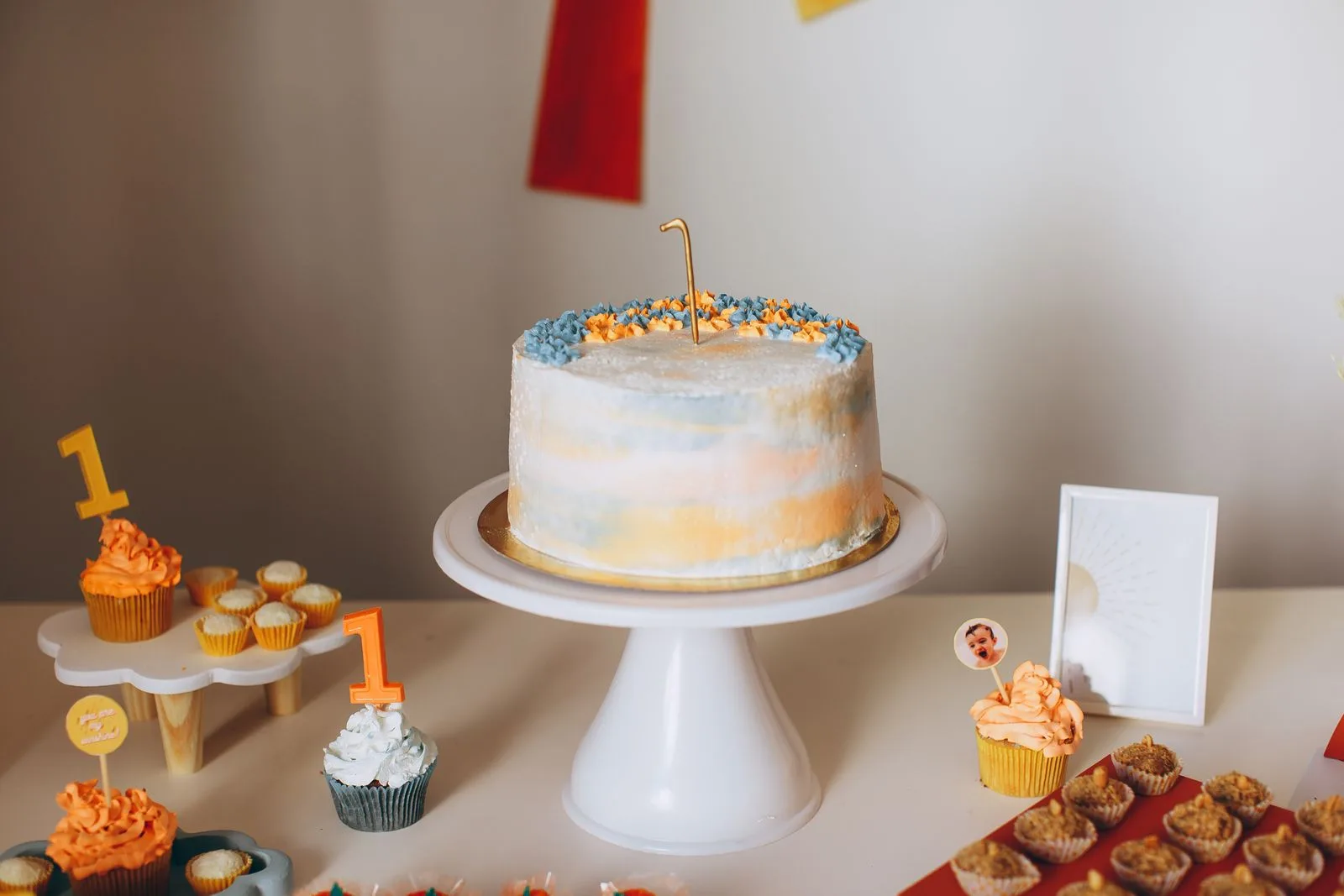 A pastel first birthday cake with a gold number one candle on a dessert table with cupcakes