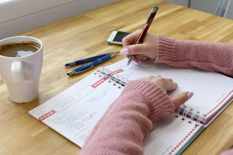 Woman writing money affirmations by hand in a spiral journal with coffee on a warm wood desk