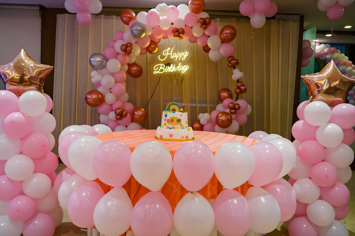 A pink and gold birthday party setup with a balloon arch, a Happy Birthday sign, and a cake