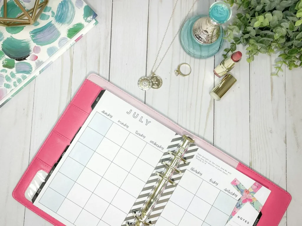 A pink ring-bound planner open to a July monthly page on a white wooden desk