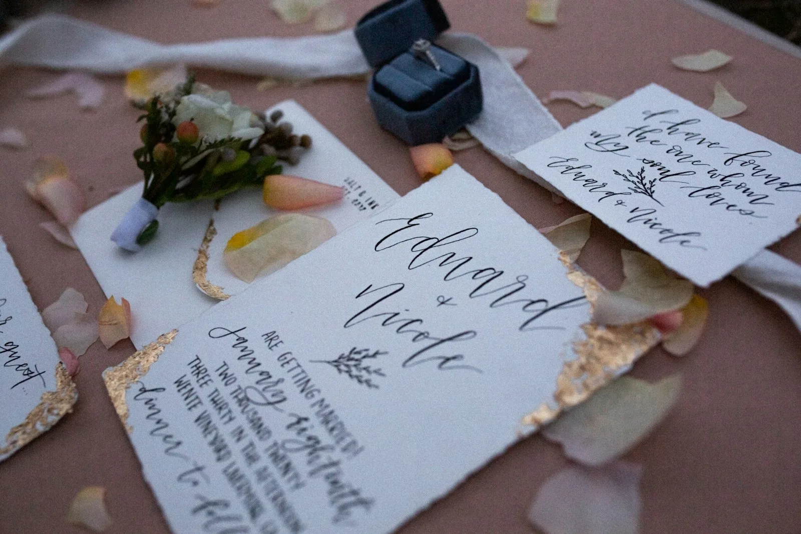 Wedding invitation suite with hand-lettered calligraphy names, a gold-edged card, petals and a ring box